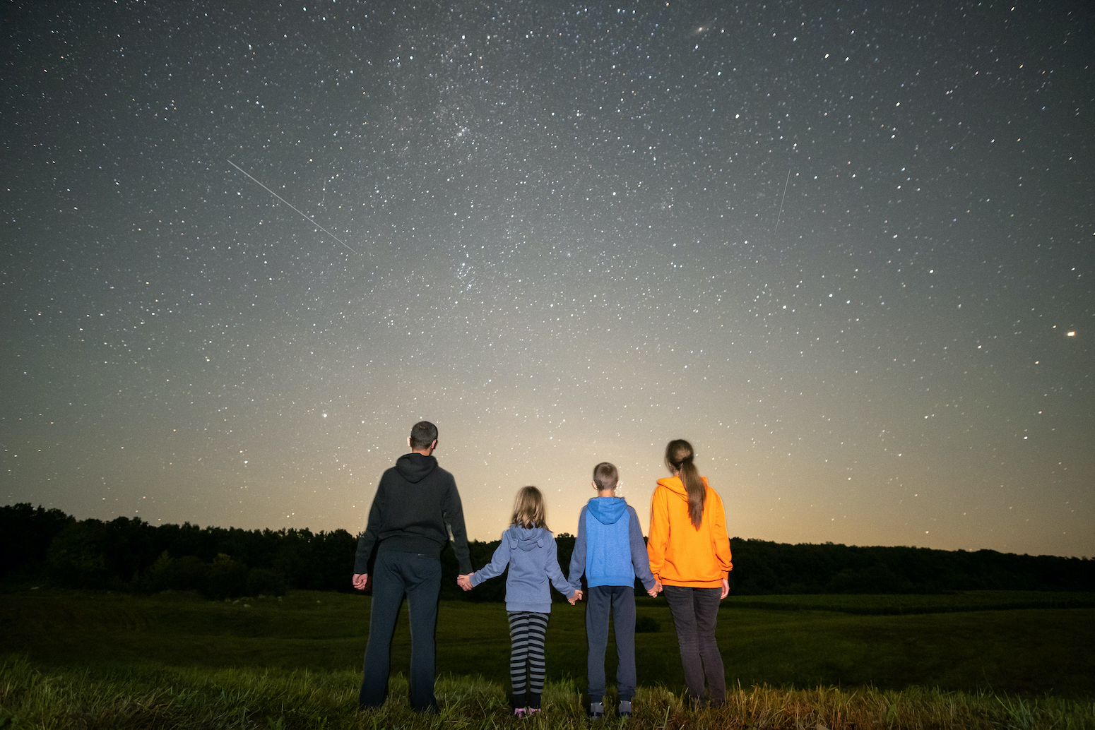Parents and their kids standing in night field observing dark sky with many bright stars.